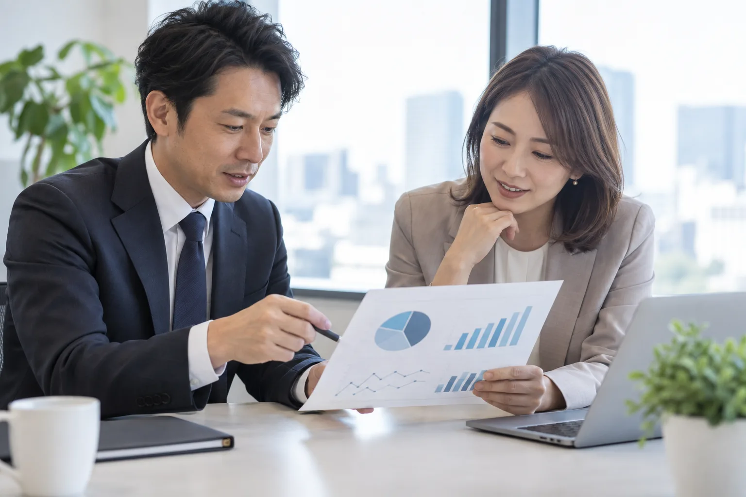 ノートパソコンの前で、グラフの載った報告書を手に内容を確かめている人